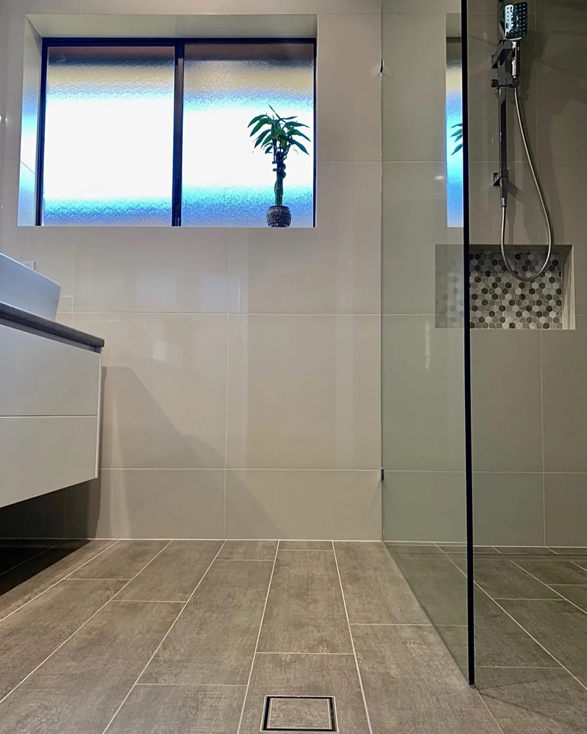 Walk-in shower with grey timber-look floor tiles falling to a square floor waste beside a frameless glass panel